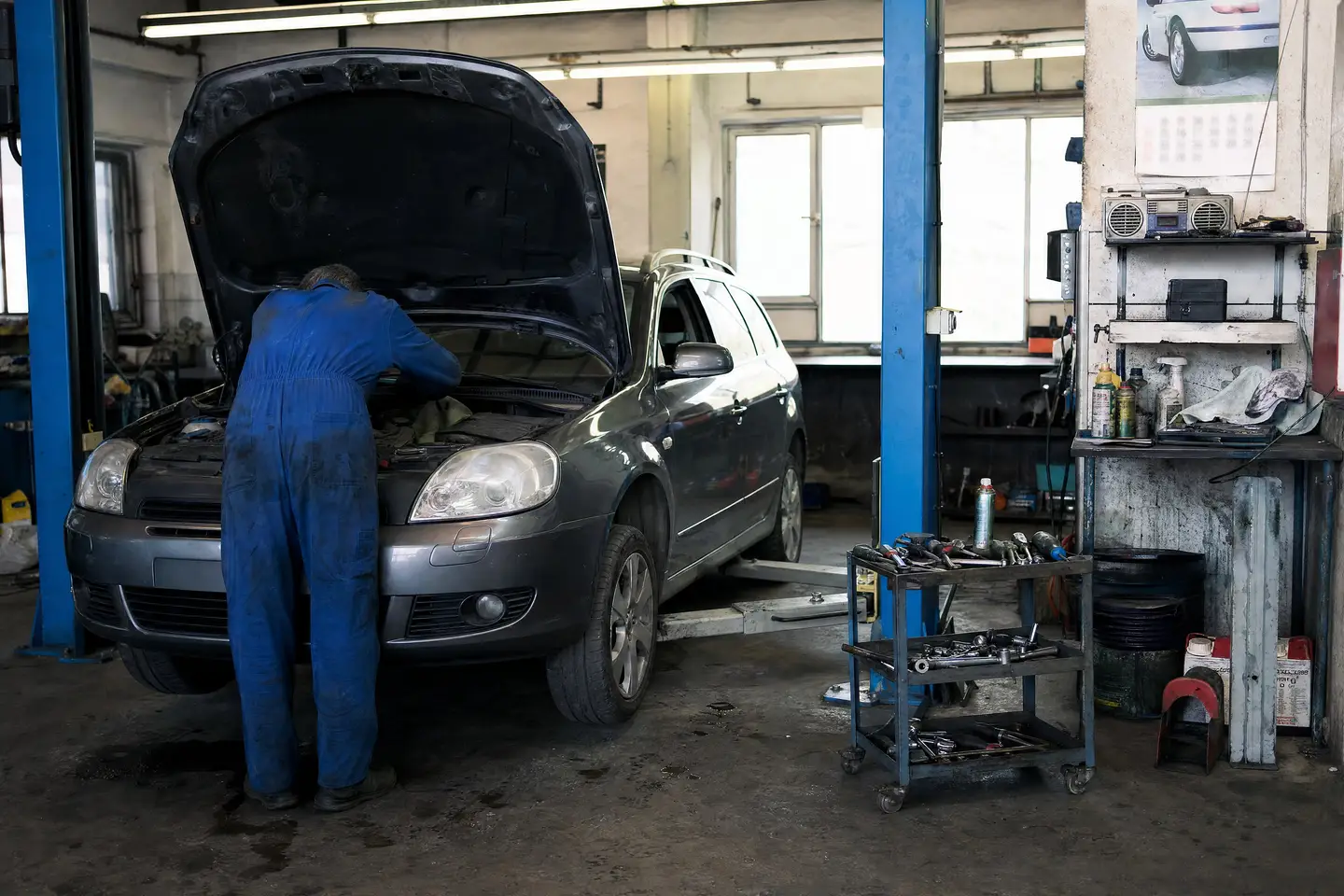Mechaniker in blauem Überkleid beugt sich in einer kleinen Garage über den Motorraum eines grauen Kombis
