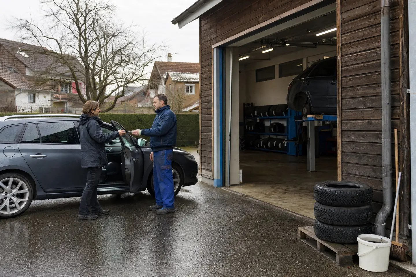 Kundin übergibt dem Mechaniker vor einer kleinen Dorfgarage mit Holzfassade den Autoschlüssel, im offenen Tor ein Auto auf der Hebebühne und Pneus im Regal