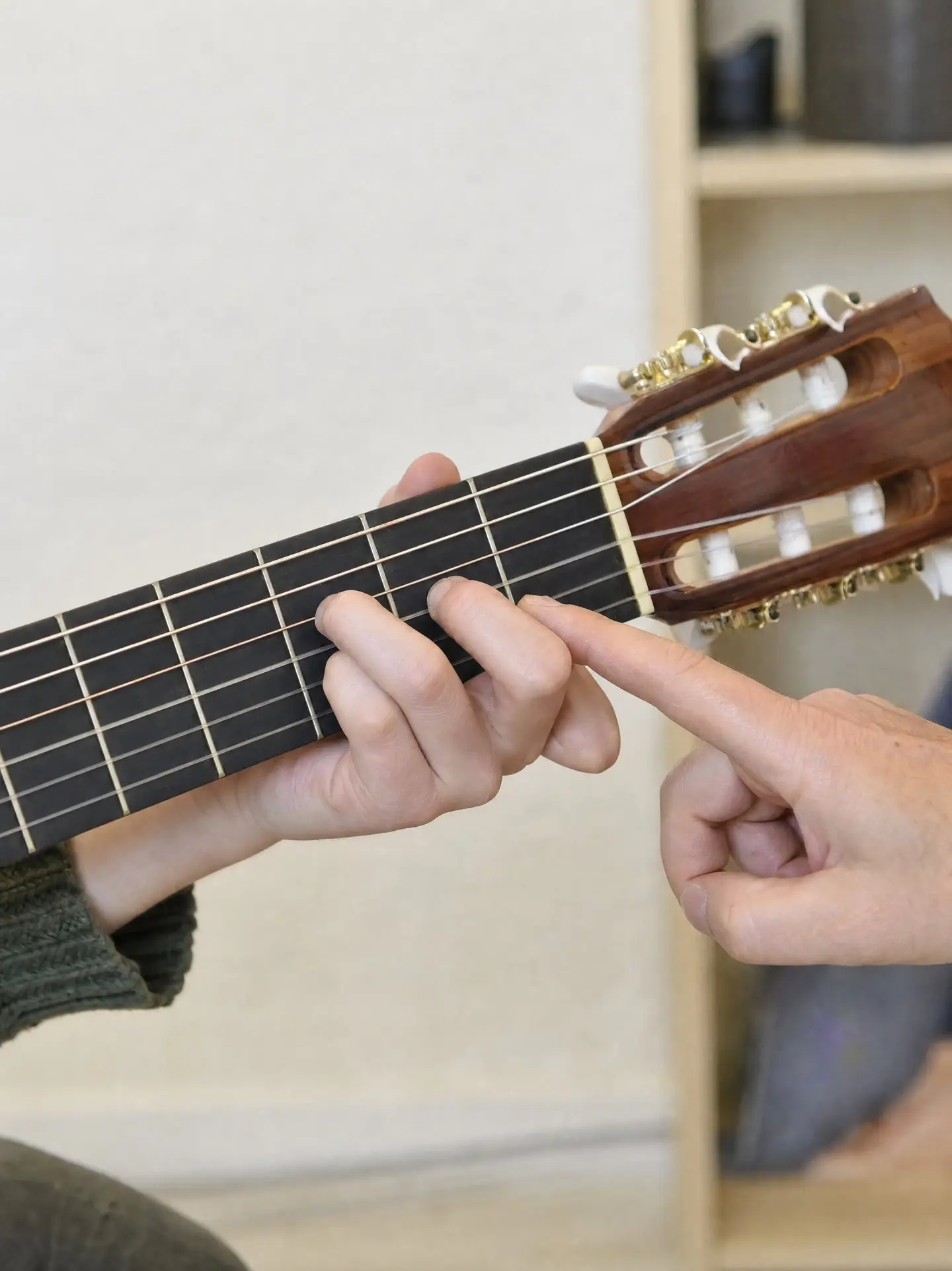 Hand greift einen Akkord auf dem Hals einer Konzertgitarre, die Lehrperson korrigiert mit dem Zeigefinger
