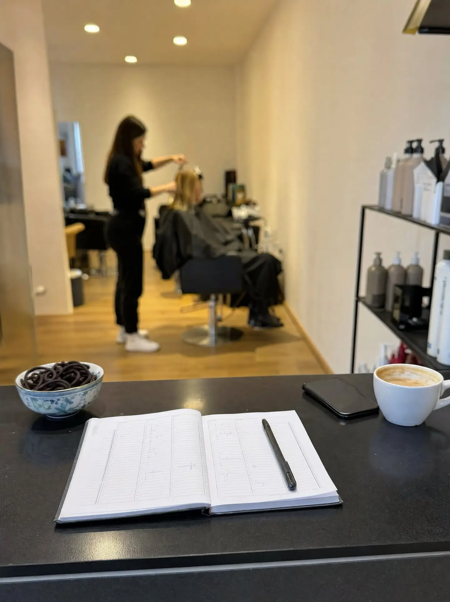 Terminbuch, Stift und umgedrehtes Handy auf der Theke eines Coiffeursalons, hinten wird einer Kundin das Haar gemacht