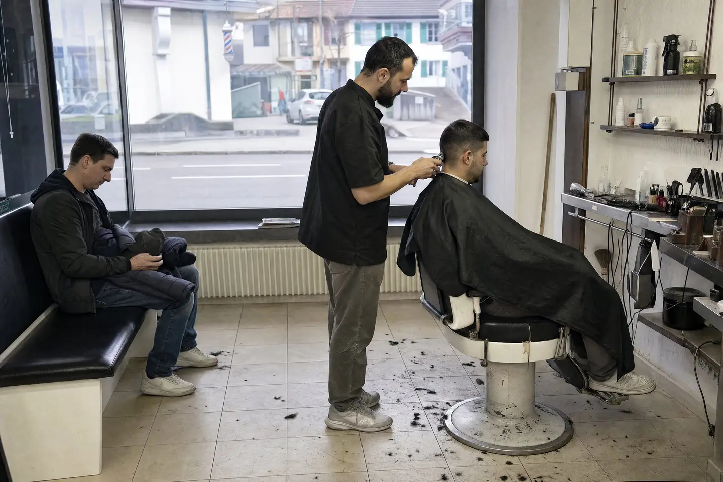 Barbier schneidet einem Kunden mit der Maschine die Haare, ein weiterer Kunde wartet auf der Bank