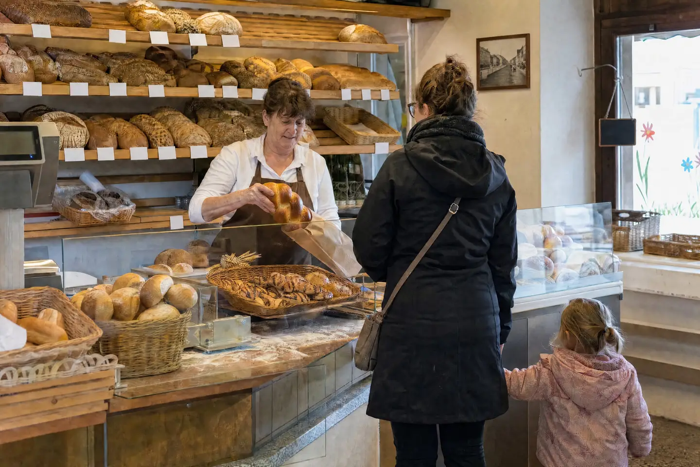 Verkäuferin in einer Dorfbäckerei packt einen Zopf ein, eine Kundin mit Kind wartet an der Theke