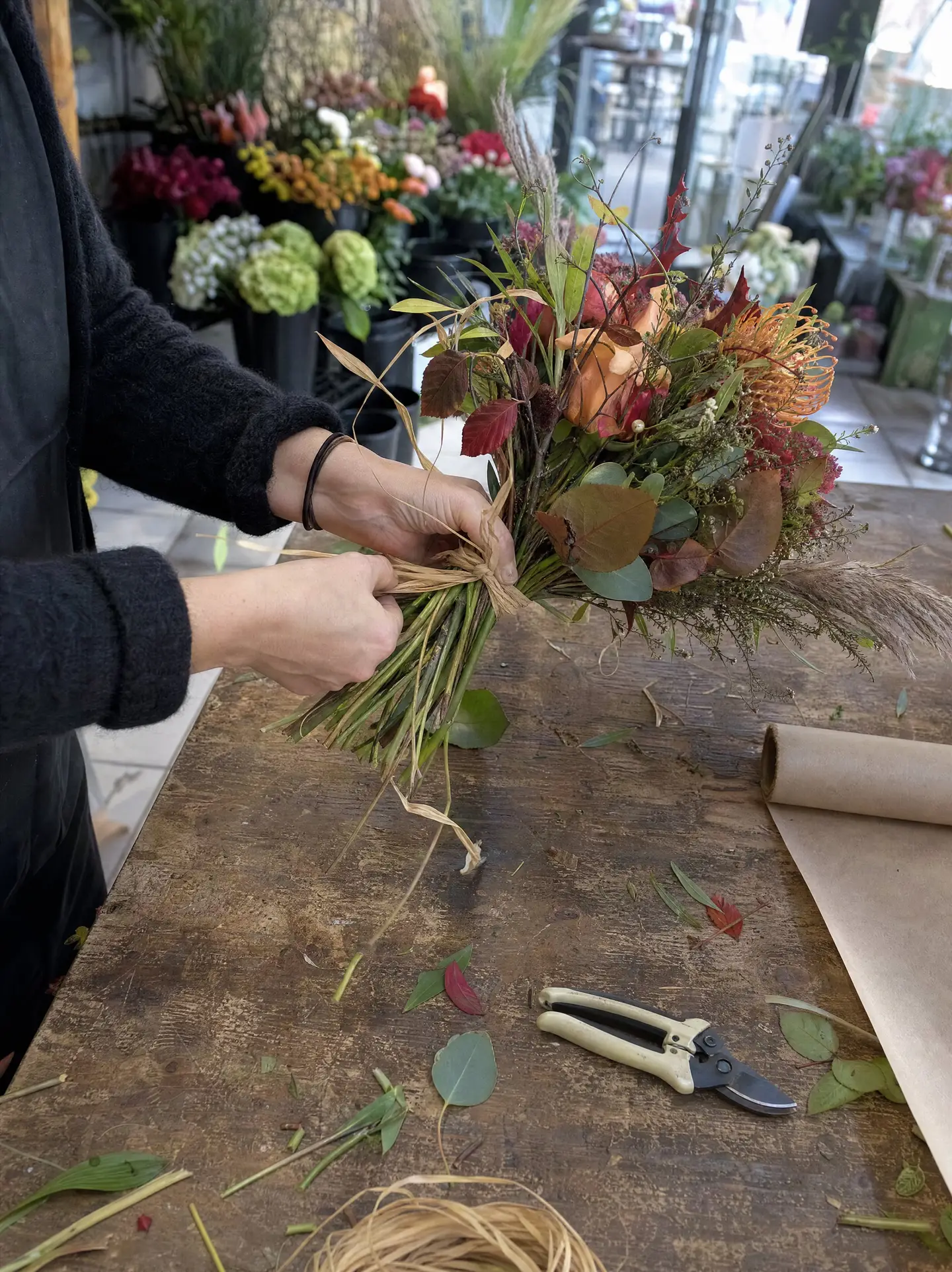 Hände einer Floristin binden einen Herbststrauss mit Bast, daneben Rosenschere und Packpapier