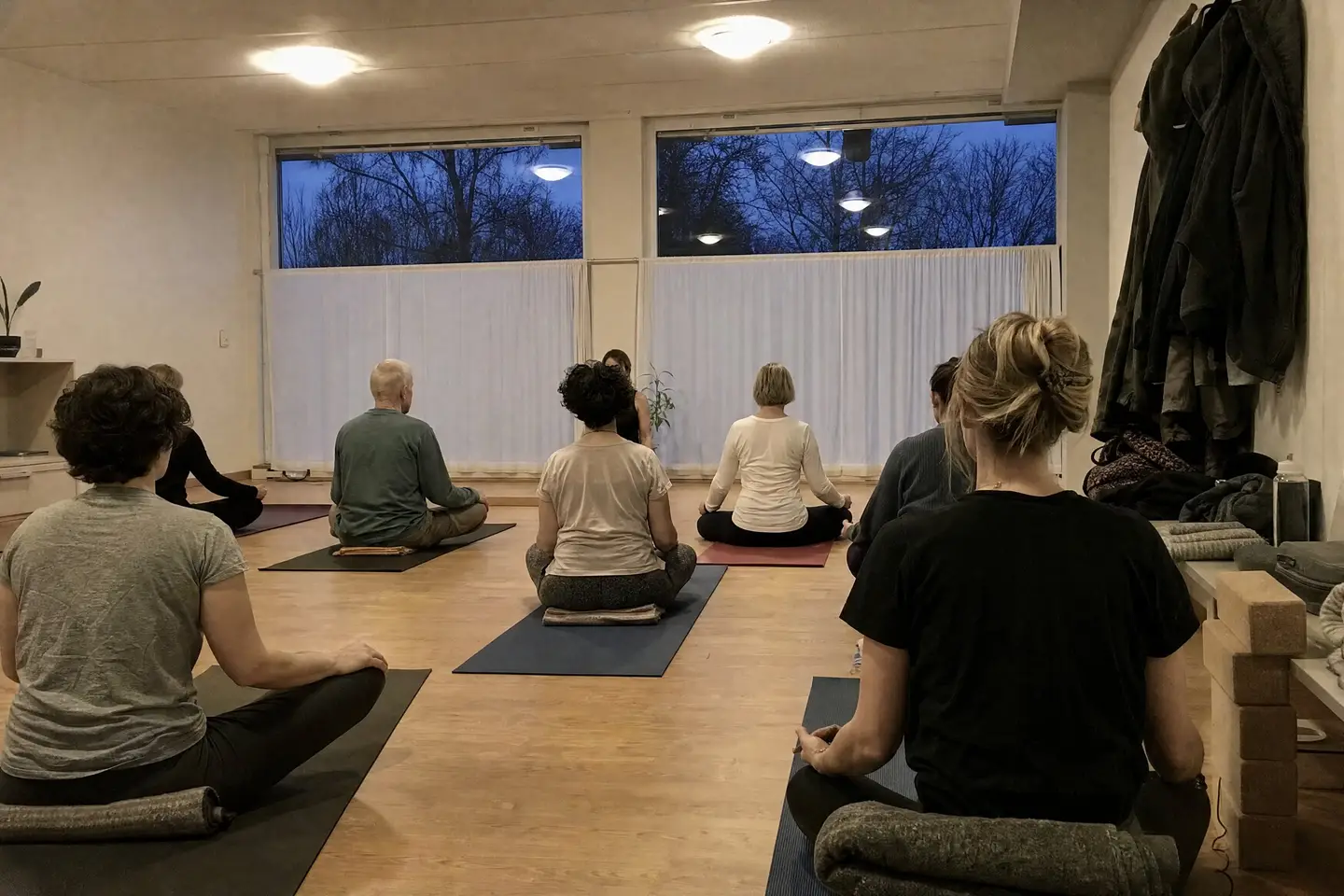 Yogakurs am Abend: Teilnehmende sitzen im Schneidersitz auf Matten, von hinten gesehen