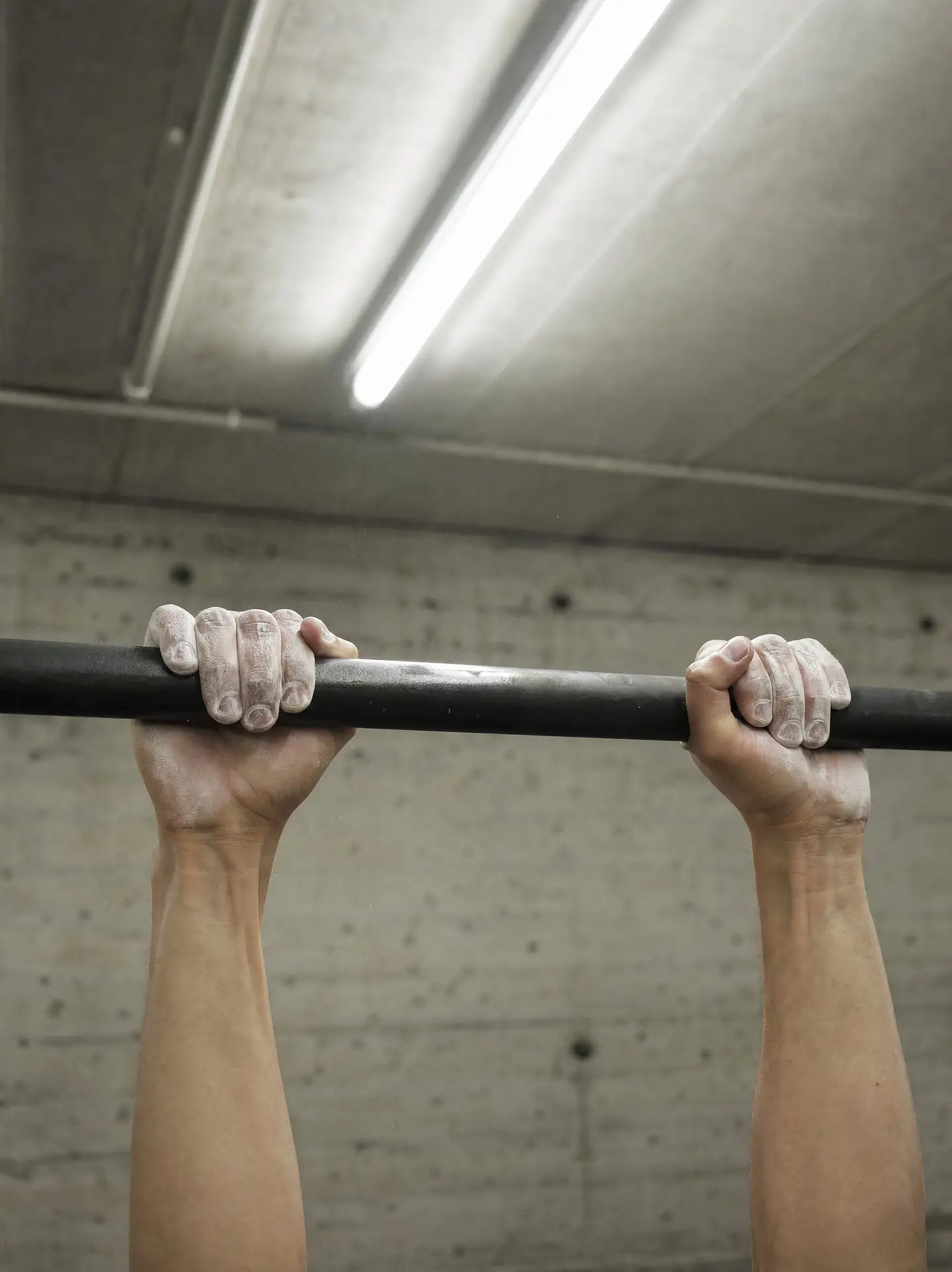 Hände mit Magnesia greifen im Fitnessraum eine Klimmzugstange, darüber eine Neonröhre