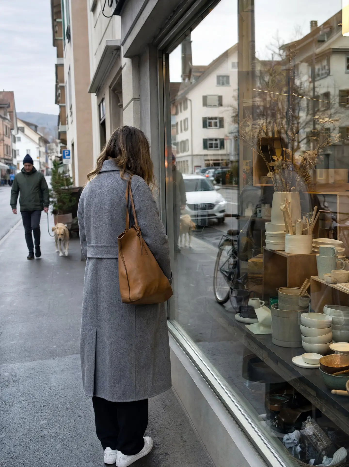 Frau im grauen Mantel bleibt auf dem Trottoir stehen und schaut ins Schaufenster eines kleinen Keramikladens