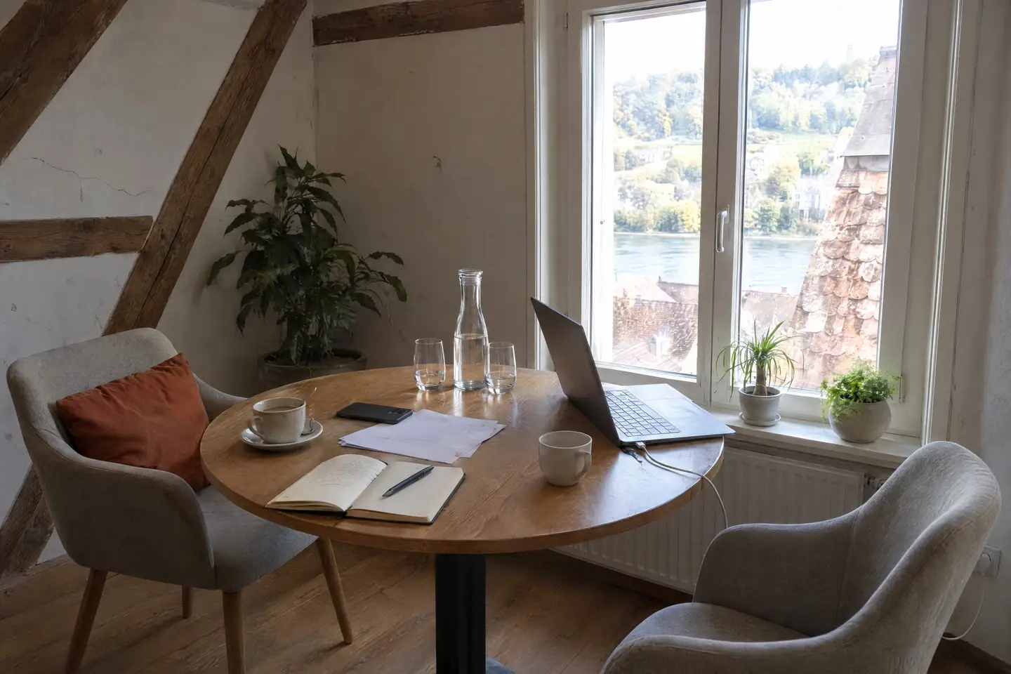 Runder Holztisch im Dachzimmer mit Kaffeetassen, Wasserkaraffe und Gläsern, offenem Notizbuch, Notizzetteln mit Handy und einem Laptop, daneben ein Sessel mit orangem Kissen, durchs Fenster Altstadtdächer und ein Fluss