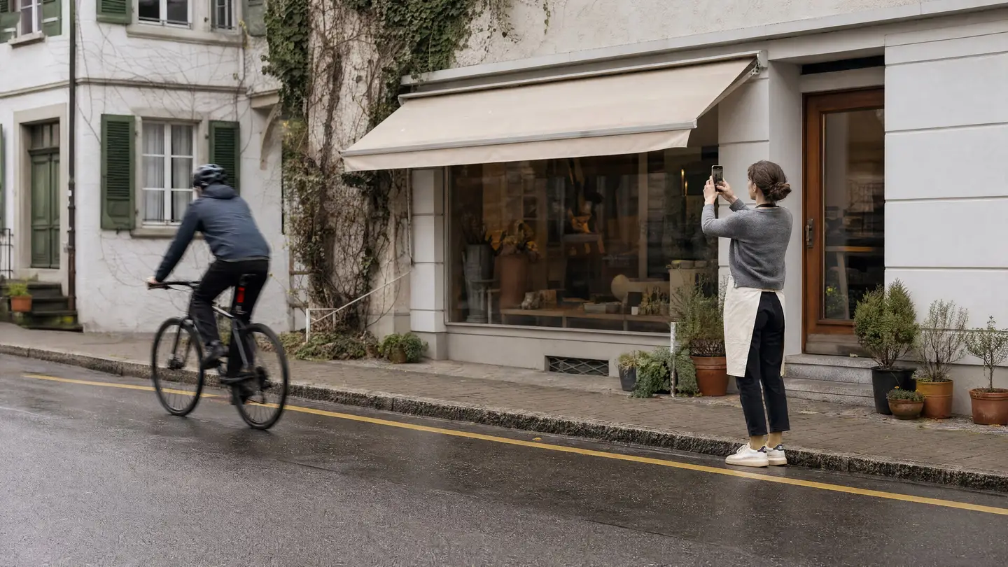 Ladeninhaberin in Schürze fotografiert mit dem Handy ihr Schaufenster, ein Velofahrer fährt vorbei