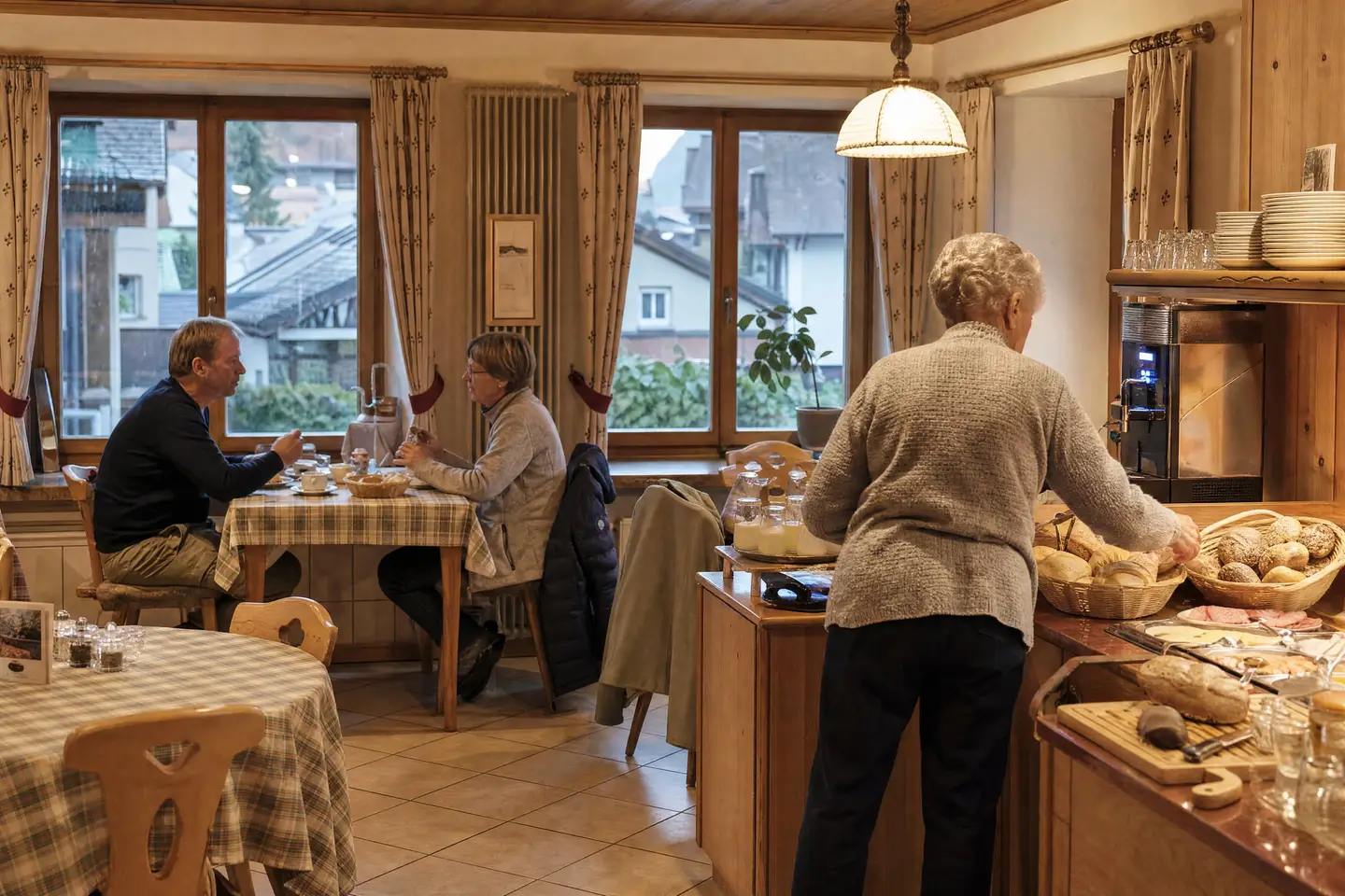 Frühstücksraum eines kleinen Familienhotels, Gäste am Fenstertisch, die Gastgeberin füllt das Brotkörbchen nach