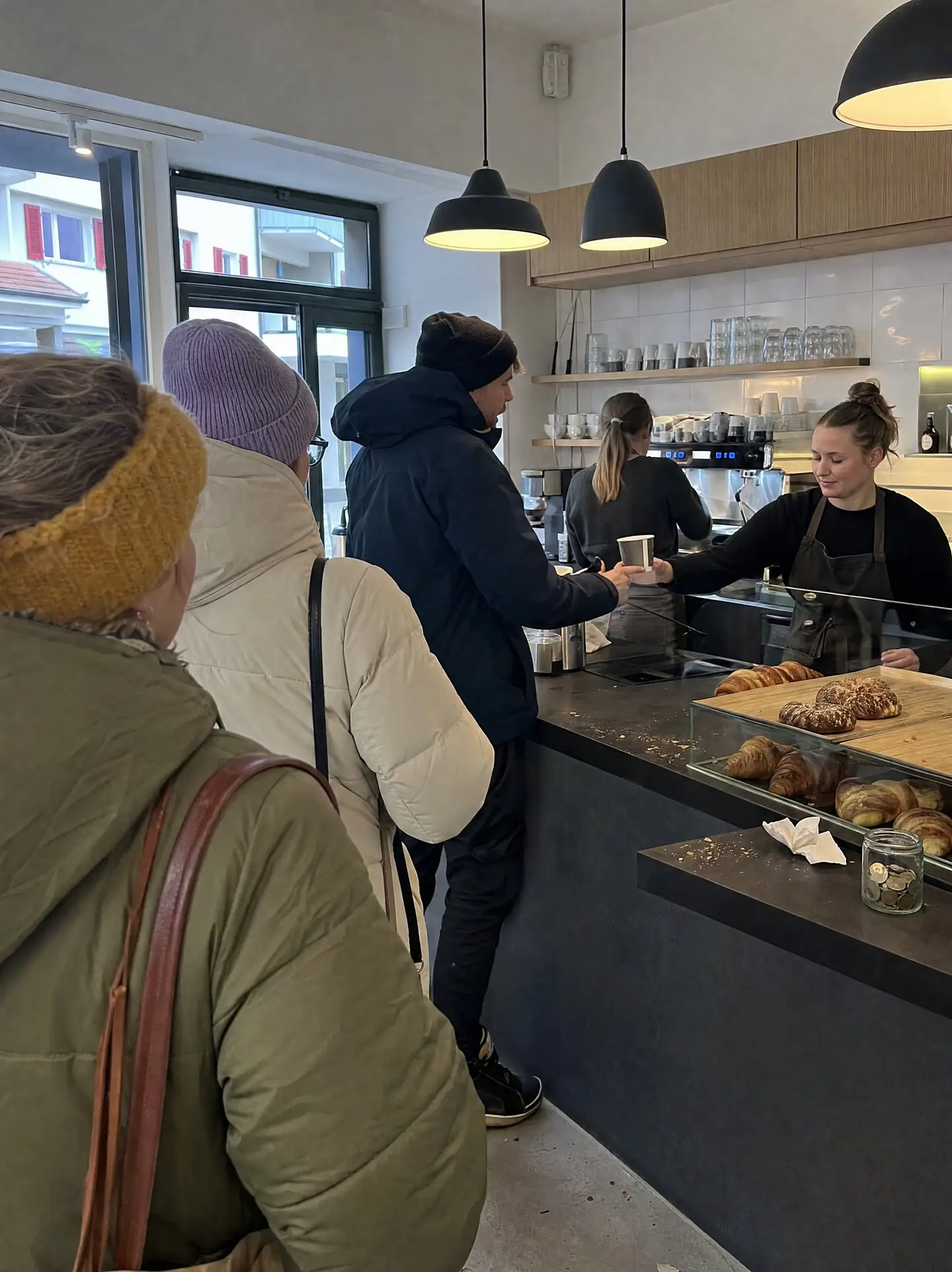 Kundschaft in Winterjacken wartet am Tresen eines kleinen Cafés, die Bedienung reicht einen Kaffeebecher hinüber