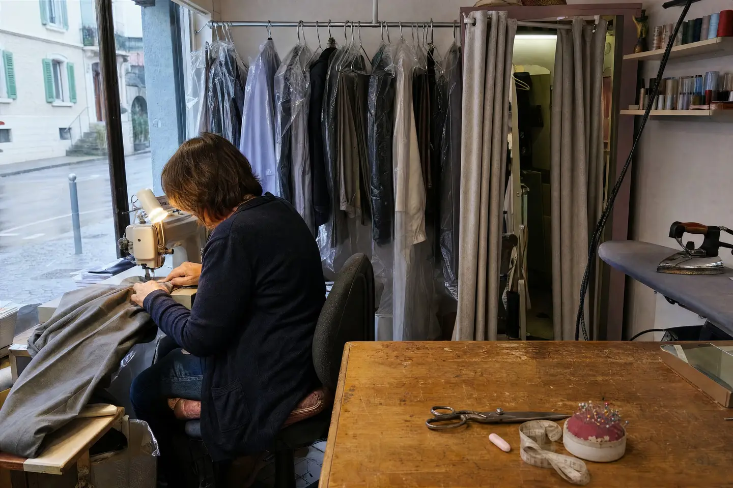 Änderungsschneiderin kürzt am Schaufenster eine graue Hose an der Nähmaschine, dahinter fertige Kleider in Schutzhüllen