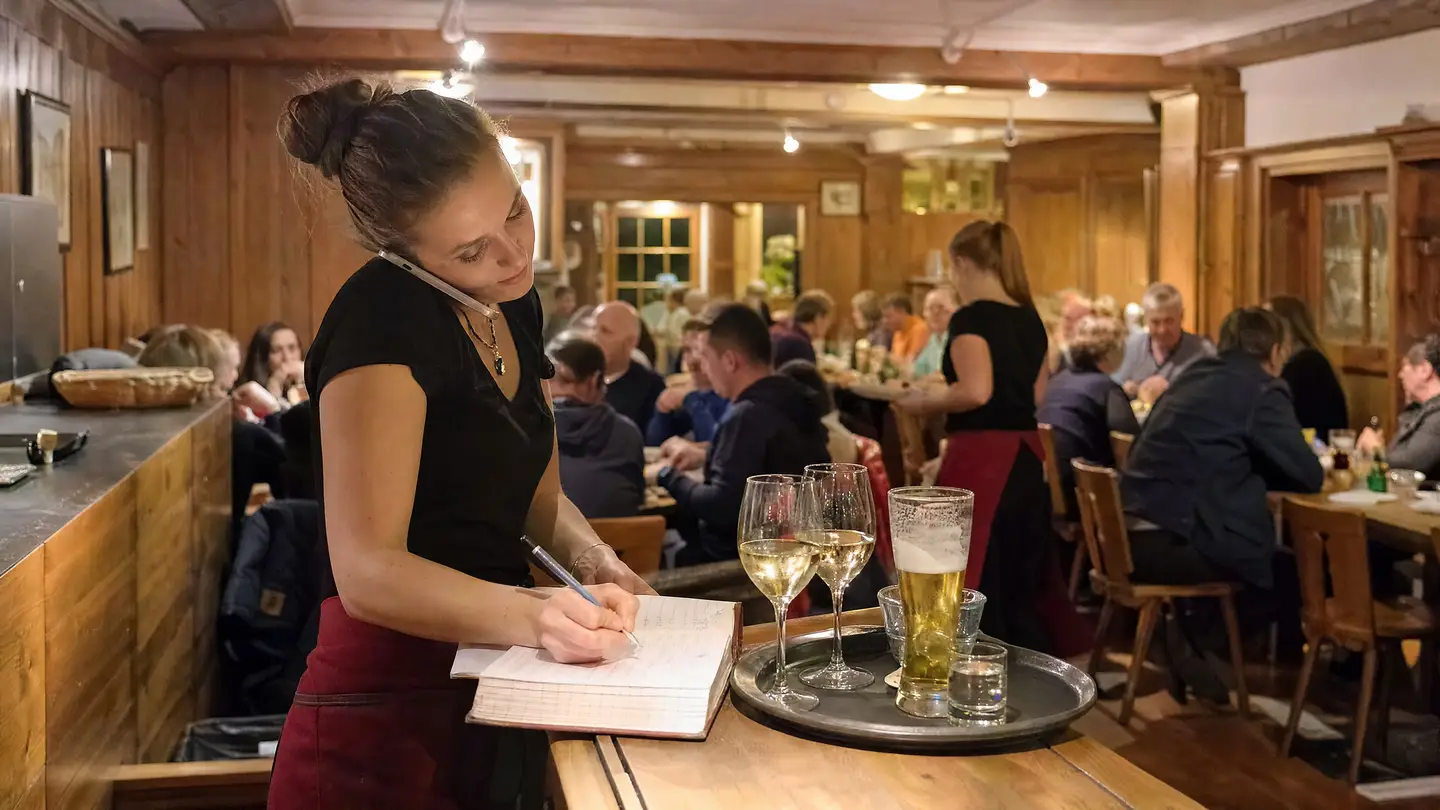 Serviceangestellte nimmt am Telefon eine Reservation entgegen und schreibt ins Buch, die Gaststube ist voll