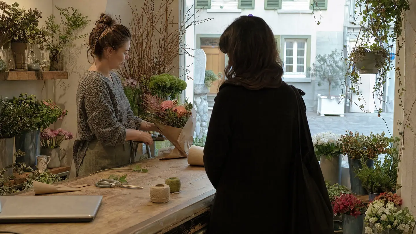Floristin bindet in ihrem Blumenladen einen Strauss, eine Kundin wartet an der Holztheke