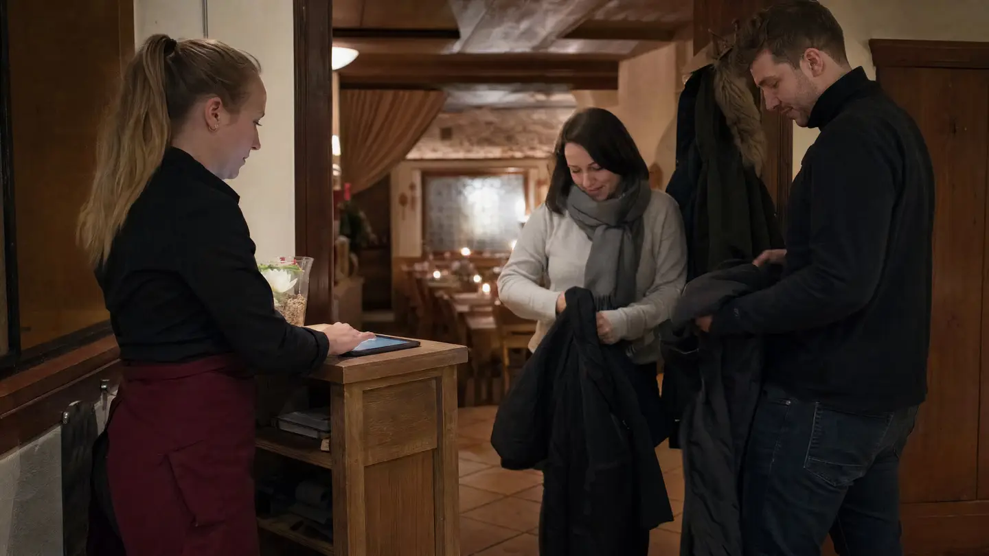Servicemitarbeiterin am Empfangspult eines Restaurants, ein Paar kommt am Abend an und zieht die Jacken aus