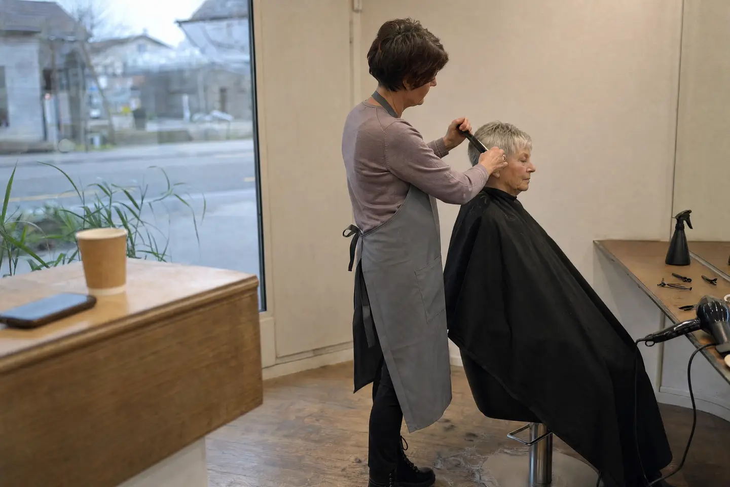 Coiffeuse arbeitet an den kurzen grauen Haaren einer älteren Kundin, vorne auf der Theke liegen ein Handy und ein Kaffeebecher