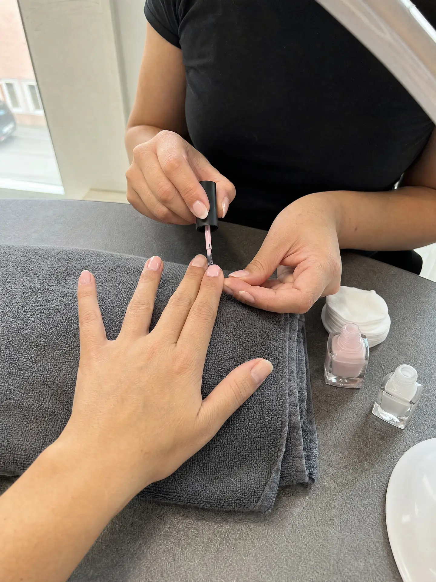 Nageldesignerin lackiert einer Kundin die Fingernägel in hellem Rosa, die Hand liegt auf einem grauen Frotteetuch