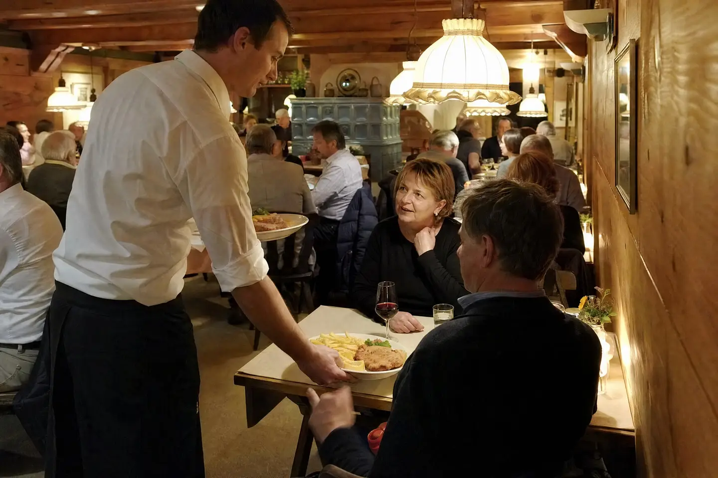 Kellner im weissen Hemd serviert in einer vollen, holzgetäferten Gaststube ein Cordon bleu mit Pommes, die Gäste am Tisch unterhalten sich